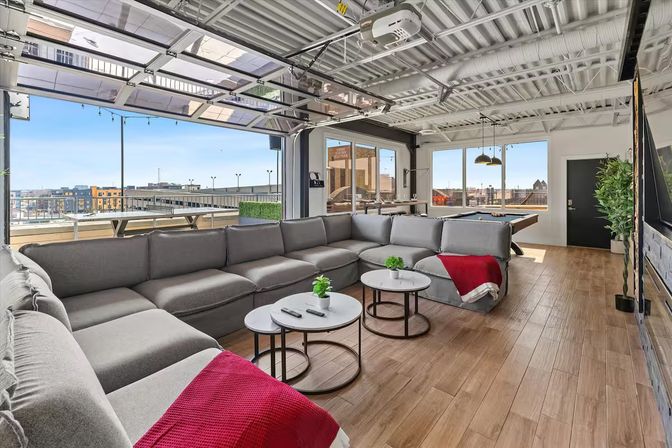 Open-plan rooftop lounge with large gray sectional sofa, round nesting coffee tables, pool table, retractable glass garage door and city skyline views