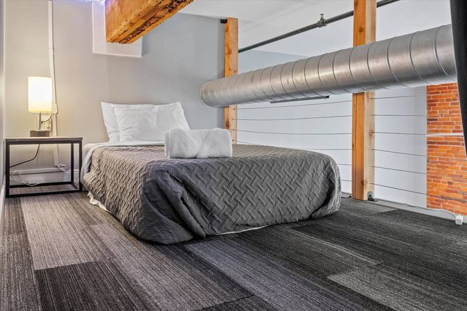 Cozy urban loft bedroom with low gray quilted bed topped with rolled white towels, exposed metal ductwork and wooden beam, nightstand lamp, carpeted floor and brick accent wall.