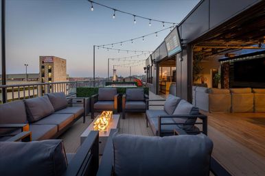 Downtown rooftop patio at dusk with modern gray sofas grouped around a long glass-topped fire table, string lights overhead, city skyline views and an open indoor lounge area.
