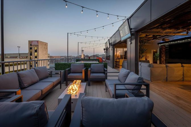 Downtown rooftop patio at dusk with modern gray sofas grouped around a long glass-topped fire table, string lights overhead, city skyline views and an open indoor lounge area.