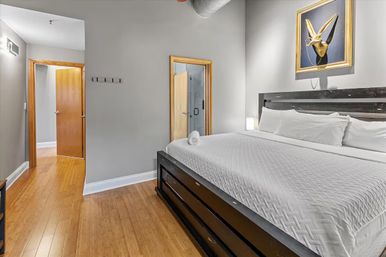 Modern bedroom with king-size bed in white linens on a dark wooden platform, hardwood floors, gray walls, playful gold rabbit wall sculpture, and open doorway to a bathroom.