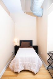 Cozy narrow loft-style bedroom with single bed in white comforter and brown pillow, black studded headboard, matching nightstands with lamp, exposed silver HVAC duct overhead, light hardwood floor and neutral walls.