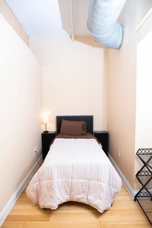 Cozy narrow loft-style bedroom with single bed in white comforter and brown pillow, black studded headboard, matching nightstands with lamp, exposed silver HVAC duct overhead, light hardwood floor and neutral walls.