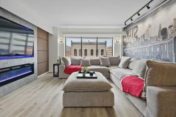 Cozy modern downtown loft living room with beige L-shaped sectional, red throws, ottoman coffee table, wall-mounted TV, linear electric fireplace, and large window overlooking a brick building.