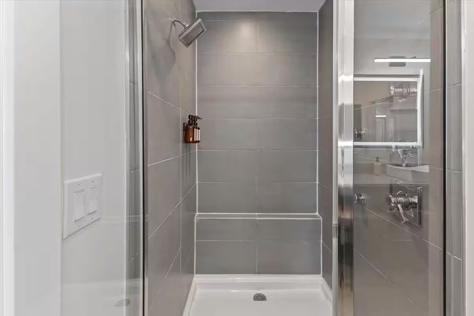 Sleek spa-like walk-in shower with large gray tiles, glass door, chrome rainfall showerhead, built-in bench and amber wall-mounted soap dispenser