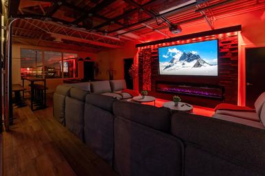 Loft-style home theater/media room with plush gray sectional facing a large wall-mounted flat-screen TV above a modern electric fireplace, red LED ambient lighting, wood accent wall, and a bar area by windows.