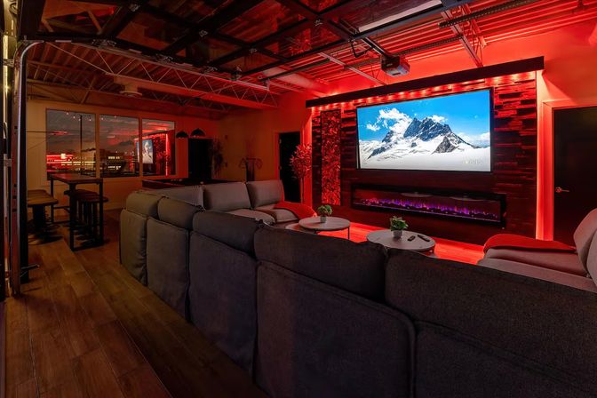 Loft-style home theater/media room with plush gray sectional facing a large wall-mounted flat-screen TV above a modern electric fireplace, red LED ambient lighting, wood accent wall, and a bar area by windows.
