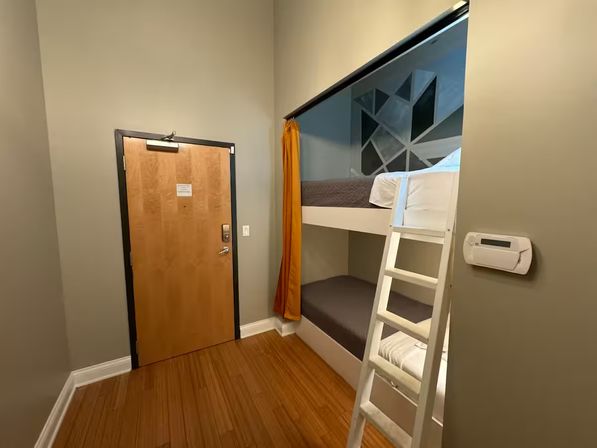 Cozy urban hostel-style room with a wooden entry door, white-framed bunk beds and ladder, mustard privacy curtain, geometric accent wall, hardwood floor, and wall thermostat.