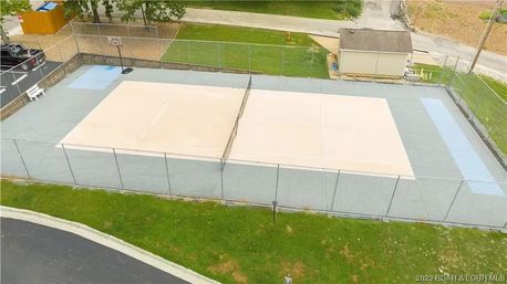 Aerial view of a fenced outdoor community sports area with two adjacent pale-tan courts separated by a central net, blue end zones, a nearby basketball hoop, storage shed, and surrounding green lawn.