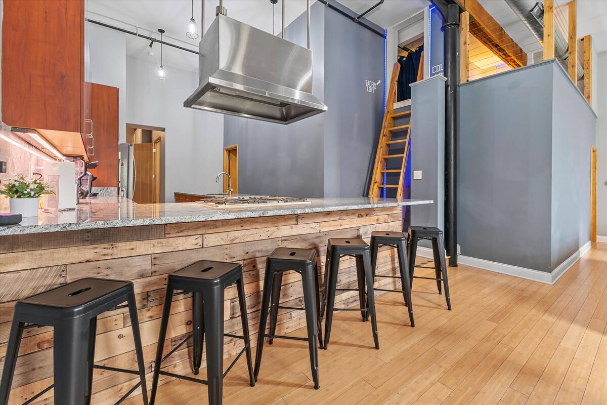 Modern loft kitchen with reclaimed wood breakfast bar, five black metal stools, granite countertop, stainless steel range hood, gray walls, ladder to an upper loft and light hardwood floors.