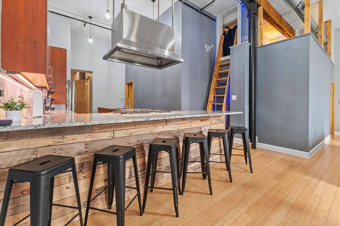 Modern loft kitchen with reclaimed wood breakfast bar, five black metal stools, granite countertop, stainless steel range hood, gray walls, ladder to an upper loft and light hardwood floors.