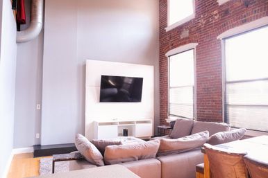 Cozy urban industrial loft living room with exposed red brick wall and tall windows, light-filled space featuring a gray sectional sofa, wall-mounted flat-screen TV, white media console, hardwood floors, and high ceilings.
