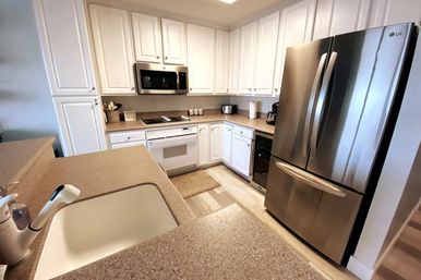 Bright modern kitchen with white cabinets, beige quartz-look countertops and breakfast bar sink, stainless-steel French-door refrigerator, built-in microwave and compact countertop appliances.