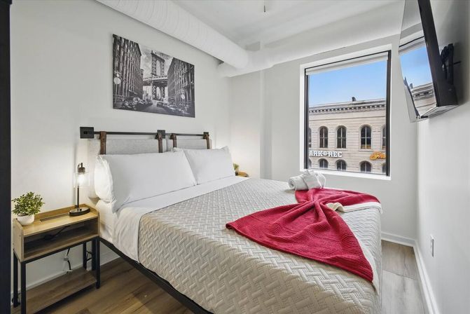 Bright modern downtown loft bedroom with queen bed in white linens, textured quilt and folded red throw, wooden nightstand with lamp and plant, black-and-white city photo above the headboard, wall-mounted TV and a window framing a neighboring stone building.