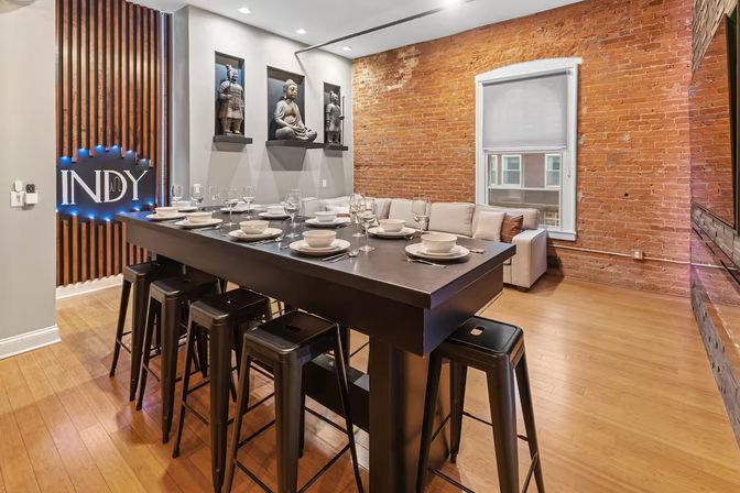 Stylish urban loft dining area with exposed brick walls and hardwood floors, long black high-top communal table set with dishes and glassware, metal bar stools, decorative Asian statues in wall niches, and a beige sofa by the window.