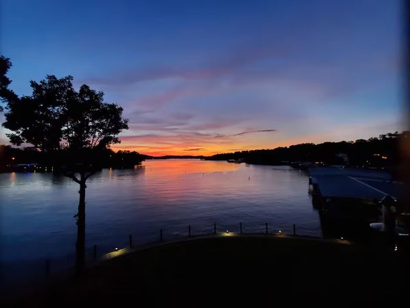 Tranquil lakeside sunset with orange and purple sky reflecting on calm water, silhouetted tree and docks along the shore.