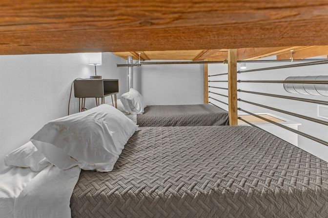 Cozy modern industrial loft sleeping area with two twin beds in gray quilted covers and white pillows, wooden overhead beam, metal horizontal railing, exposed ductwork, and a small bedside table with lamp.