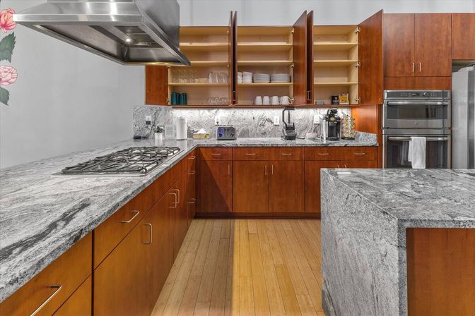 Spacious modern kitchen with gray granite countertops and island, warm wood cabinets, gas cooktop under stainless steel hood, double wall ovens and open upper shelves with dishes on light wood floors.