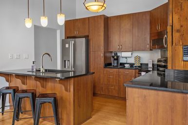 Sleek modern kitchen with warm wood cabinets, black granite countertops, stainless steel refrigerator, island sink with three metal bar stools and hanging pendant lights.