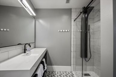 Sleek modern gray bathroom with white floating vanity, black faucet, large mirror, geometric floor tiles and glass-enclosed marble-tile shower with black rain panel
