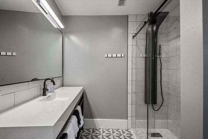 Sleek modern gray bathroom with white floating vanity, black faucet, large mirror, geometric floor tiles and glass-enclosed marble-tile shower with black rain panel
