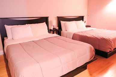 Bright hotel-style guest room with two plush double beds, dark wood headboards, white pillows, pink comforters, bedside lamp on a small table and hardwood flooring.