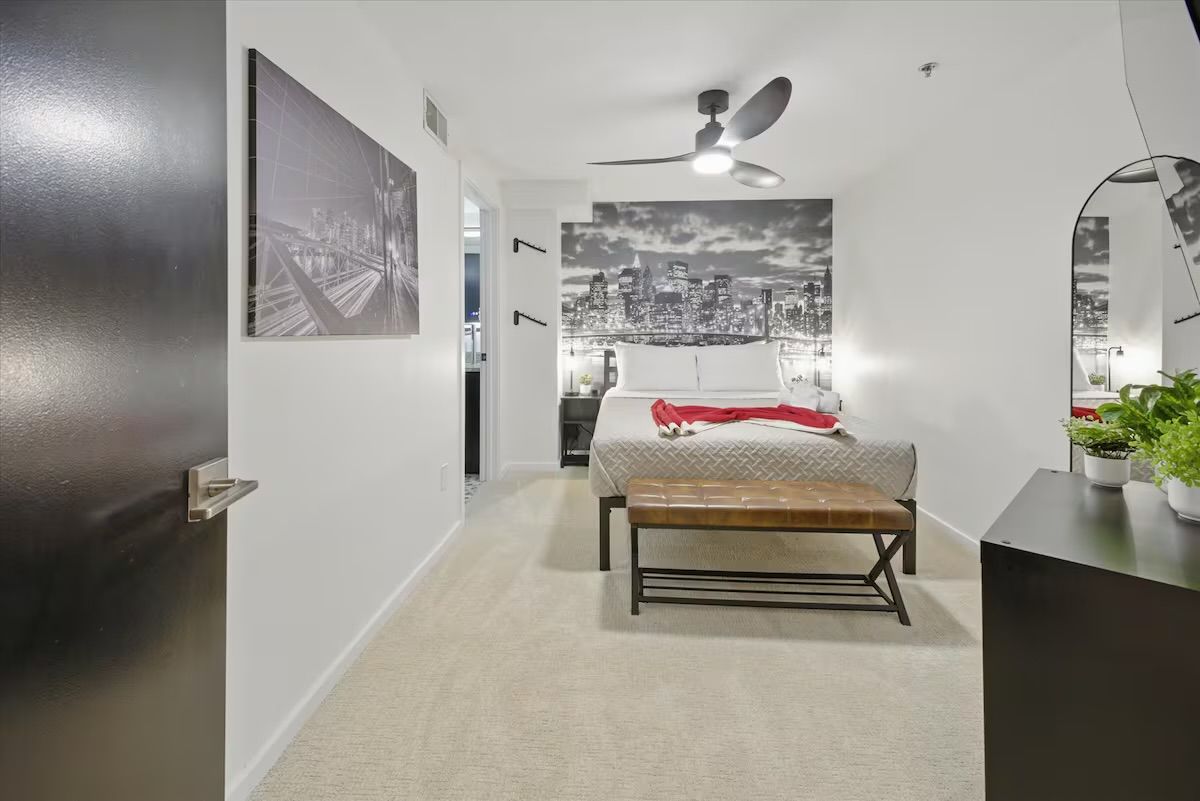 Bright modern apartment bedroom with queen bed, red throw, leather bench, ceiling fan and black-and-white city skyline mural