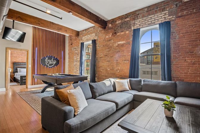Cozy industrial-style Indianapolis loft living room with exposed brick walls, wooden ceiling beams, gray sectional sofa, pool table and arched windows letting in city light