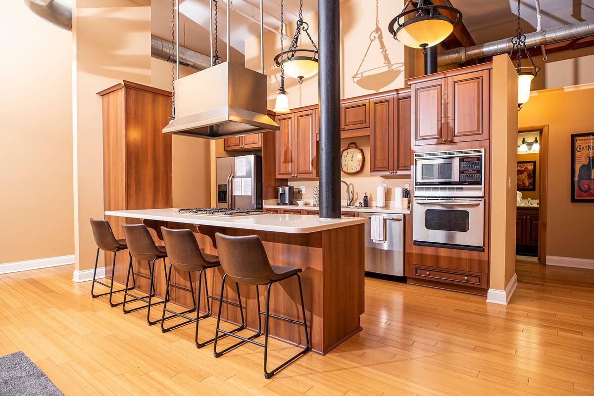 Modern urban loft kitchen with large wooden island and five bar stools, stainless steel appliances, hanging pendant lights, exposed ductwork and hardwood floors.