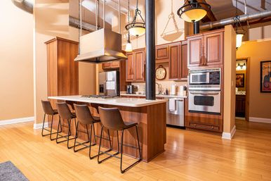 Modern urban loft kitchen with large wooden island and five bar stools, stainless steel appliances, hanging pendant lights, exposed ductwork and hardwood floors.