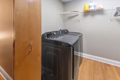Modern home laundry room with matching dark gray washer and dryer, wire shelf holding detergent jugs and hangers, light gray walls, hardwood floor, and a partially open wooden door.