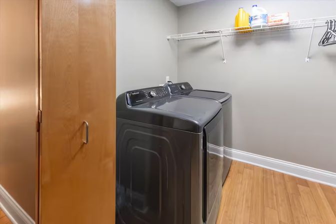 Modern home laundry room with matching dark gray washer and dryer, wire shelf holding detergent jugs and hangers, light gray walls, hardwood floor, and a partially open wooden door.
