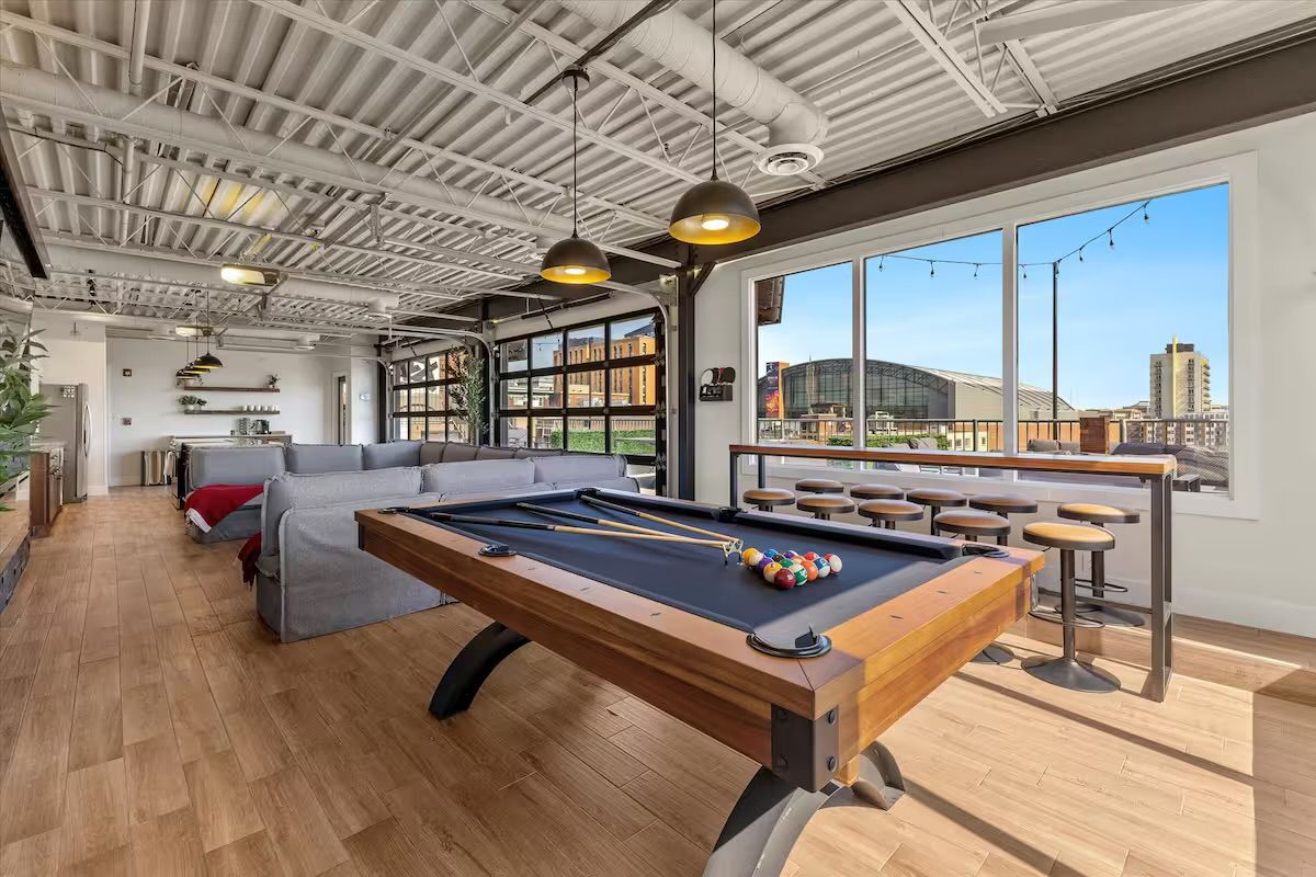 Bright industrial-style loft game room with a wooden pool table and racked colorful balls, gray sectional sofa, bar-height stools along large windows overlooking a city skyline, pendant lights, exposed ceiling, and hardwood floors.