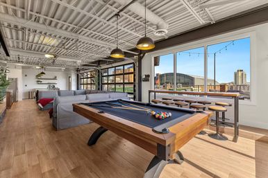 Bright industrial-style loft game room with a wooden pool table and racked colorful balls, gray sectional sofa, bar-height stools along large windows overlooking a city skyline, pendant lights, exposed ceiling, and hardwood floors.
