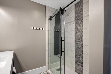Modern bathroom with glass sliding shower door, marble-look subway tiles and vertical gray mosaic accent strip, matte-black shower panel and hardware, light gray walls and white vanity edge.