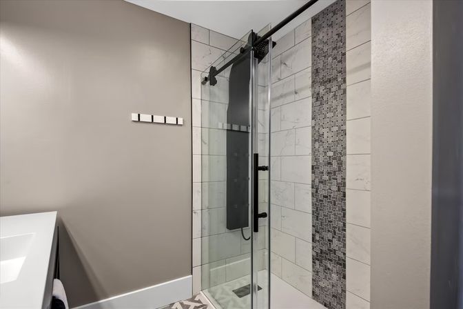 Modern bathroom with glass sliding shower door, marble-look subway tiles and vertical gray mosaic accent strip, matte-black shower panel and hardware, light gray walls and white vanity edge.