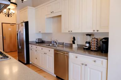 Bright white residential kitchen with shaker cabinets, stainless steel refrigerator and dishwasher, double sink and gray countertop, toaster, blender, knife block and single-serve coffee maker with K‑cup carousel on hardwood floors.