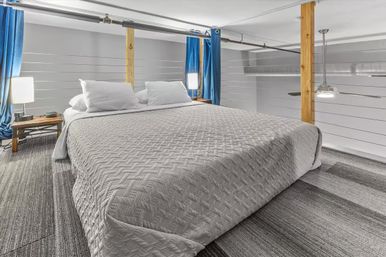 Cozy urban loft bedroom with low ceiling, large bed in a textured gray quilt, white pillows, wooden support posts, blue curtains, bedside lamps, striped carpet and exposed ductwork