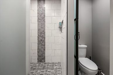 Sleek residential gray bathroom interior with glass shower door, walk-in shower lined with large white tiles and a vertical dark mosaic accent, hexagonal gray floor tiles, and a white toilet in an adjacent stall.