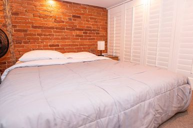 Inviting bedroom with large bed in a light-gray comforter, exposed red brick accent wall, white plantation shutters and a wooden nightstand with bedside lamp.