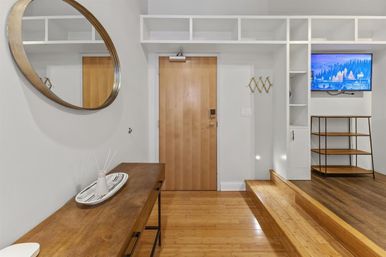 Modern apartment entryway with light wood door, round wall mirror, wooden console table, built-in white cubbies, wall-mounted TV and hardwood floors.