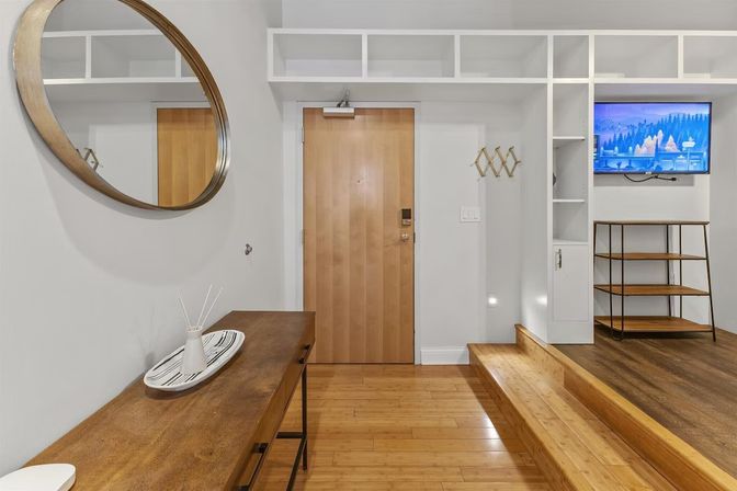 Modern apartment entryway with light wood door, round wall mirror, wooden console table, built-in white cubbies, wall-mounted TV and hardwood floors.