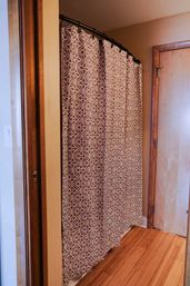 Geometric gray-and-white shower curtain on a curved black rod in a cozy wood-trimmed bathroom with light hardwood floor and a wooden door.