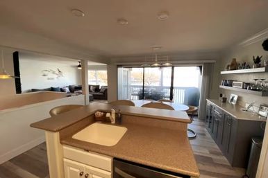 Bright open-plan kitchen and dining area with a peninsula sink and bar stools, round dining table, sliding glass doors to a balcony overlooking water, adjacent living room with sectional sofa and decorative shelving