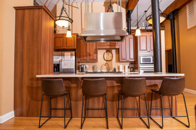 Open-concept loft kitchen with warm wood cabinets, large island and quartz countertop, stainless steel range hood, four brown bar stools, pendant lights, exposed beams and hardwood floors.