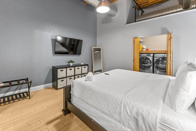 Cozy modern bedroom with gray walls, hardwood floor, queen bed with white linens, wall-mounted TV, cube storage, full-length mirror and in-room washer and dryer visible behind open doors
