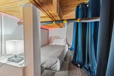 Cozy compact loft bedroom nook with single bed and quilted gray bedding, wooden ceiling beams, carpeted floor, blue privacy curtains, bedside lamp and red LED accent lighting.