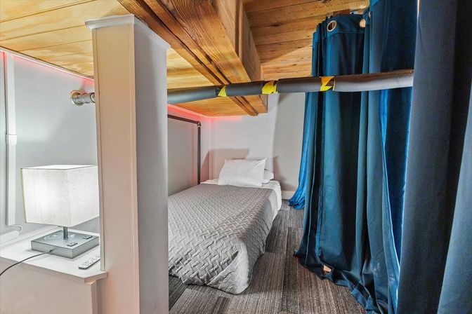 Cozy compact loft bedroom nook with single bed and quilted gray bedding, wooden ceiling beams, carpeted floor, blue privacy curtains, bedside lamp and red LED accent lighting.