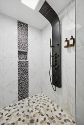 Modern spa-like shower with black curved rainfall shower panel, white marble wall tiles, vertical black mosaic tile niche, pebble-pattern floor, and amber soap dispensers