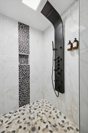 Modern spa-like shower with black curved rainfall shower panel, white marble wall tiles, vertical black mosaic tile niche, pebble-pattern floor, and amber soap dispensers